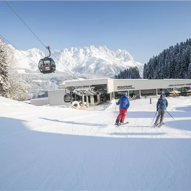 A winter landscape with snow-covered mountains in the background. Two skiers enjoy the slopes near a gondola lift.