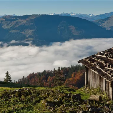 A beautiful mountain landscape with an old cabin in the foreground. Below the mountain, we see a blanket of clouds.