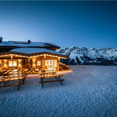 A cozy mountain cabin in the snow, brightly lit at dusk. In the background, impressive mountains can be seen.