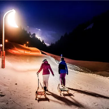 Two people are pulling sleds on a lit snow slope at night. In the background, the lights of the mountain are shining.