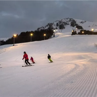 A group of skiers is skiing on a compressed snow slope. In the background, a wooded hill and a dark sky are visible.