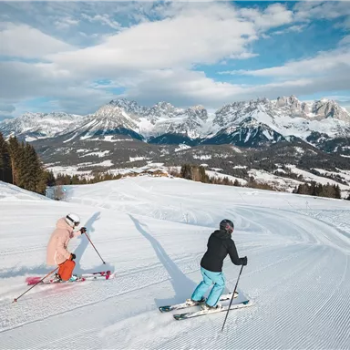 Two skiers are skiing on a snowy slope. In the background, impressive mountains and a blue sky are visible.