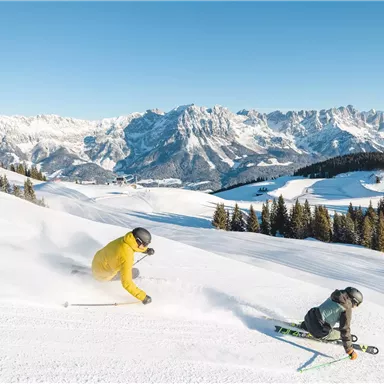 Two skiers are skiing over fresh snow in the mountains. In the background, majestic mountains and a clear blue sky can be seen.