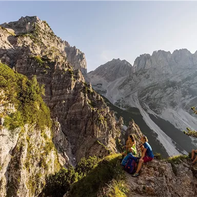 A stunning mountain landscape with steep rocks and green forests. Two hikers enjoy the view of the surrounding peaks.