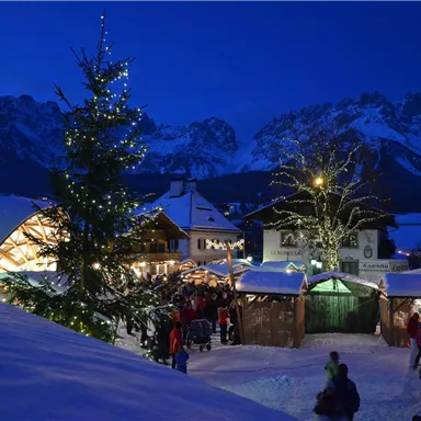 A festive Christmas market in a snowy mountain landscape. The stalls are illuminated and there is a decorated Christmas tree.
