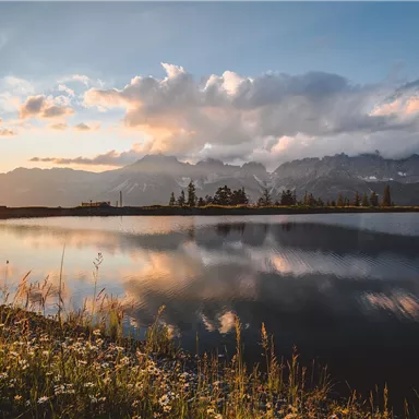 A tranquil landscape with a lake that reflects the mountains. The sun sets behind the mountains, bathing the scene in warm light.