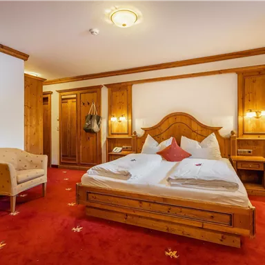 A cozy bedroom with wooden furniture and a double bed. The floor is covered with a red carpet and there is a comfortable armchair.