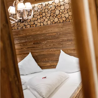 A cozy bedroom with a wooden bed and white pillows. The walls are decorated with wooden logs, and an elegant lamp hangs from the ceiling.