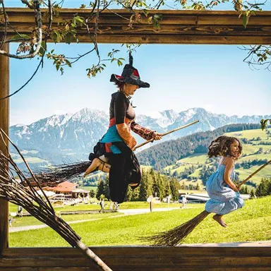 Ein Mädchen und eine Hexe fliegen fröhlich auf Besen in einem malerischen Landschaftsrahmen. Im Hintergrund sind grüne Hügel und Berge sichtbar.