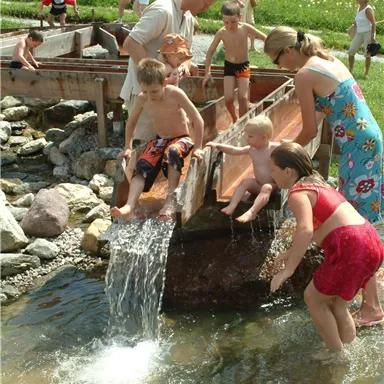 A group of children and adults are playing by the water. They are using a small slide to glide into the water and are having fun.