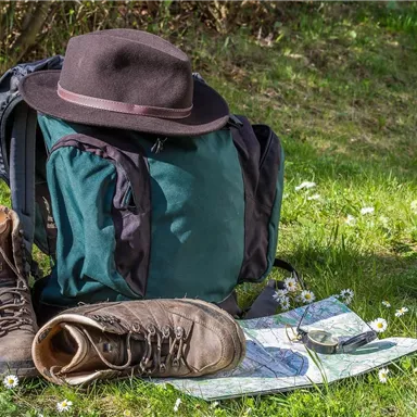 A backpack is standing on a meadow, next to a pair of hiking boots and a map. A hat lies on the backpack and in the background, fresh grass and small flowers can be seen.