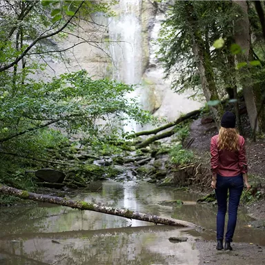 A woman stands at a small waterfall in a forested area. The forest is green and lush, with a clear water pool in the foreground.