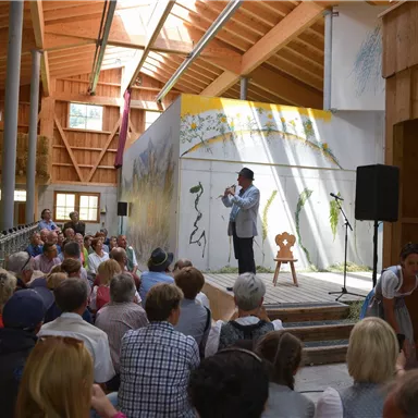 A speaker stands on stage and speaks to an audience in the background. The event space has bright wooden architecture and artistic wall design.