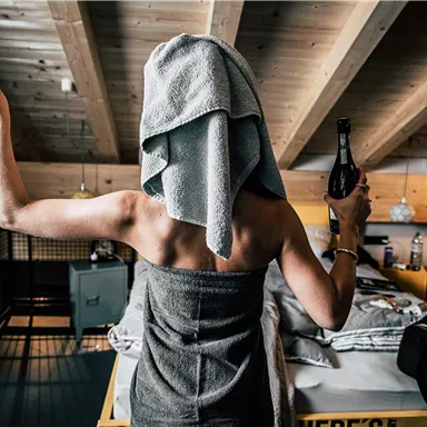 A person with a towel on their head is holding a glass in one hand and a bottle in the other. The room has a cozy atmosphere with wooden details and a modern decor.