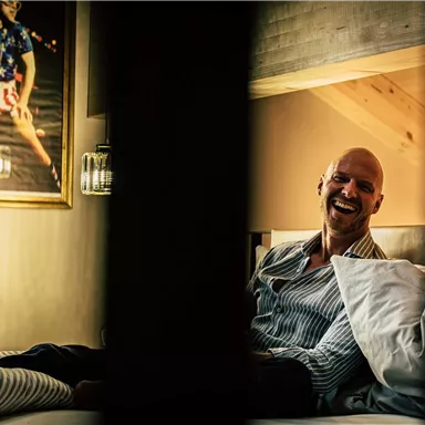 A smiling man is sitting on a bed in a cozy room. In the background, a lively painting is hanging on the wall.