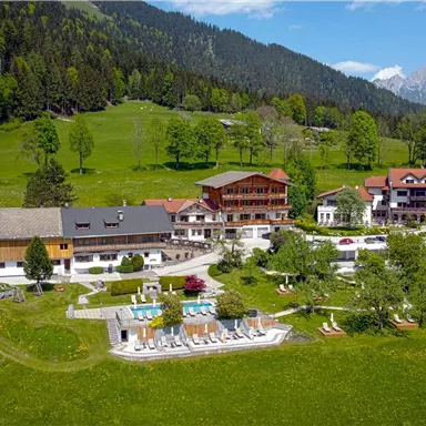 A picturesque landscape with a hotel complex, surrounded by forests and mountains. In the foreground, there is a pool area and green meadows.