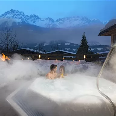 A romantic outdoor hot tub surrounded by mist and mountains in the background. The scene is illuminated in the evening with soft light.
