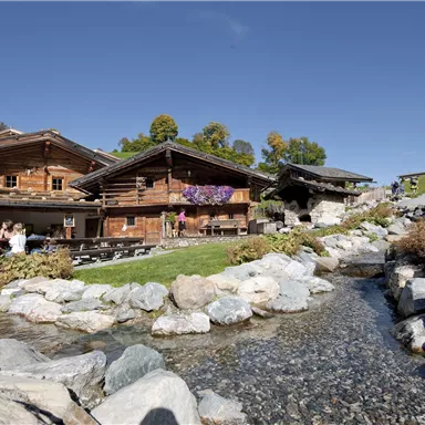 A picturesque alpine hut with wooden facades and colorful flowers. In the foreground, a clear stream flows over smooth stones.
