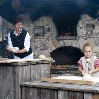 A woman and a child are standing at wooden tables and preparing dough. In the background, a traditional stone oven can be seen.