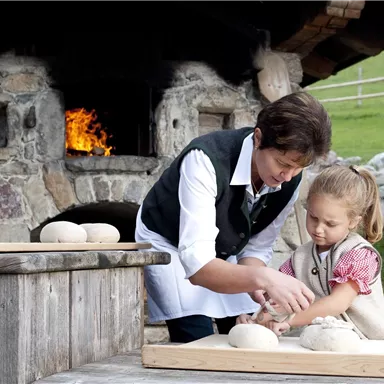 A woman and a little girl are baking bread together in front of an open fireplace. Both are working at a wooden table with dough.