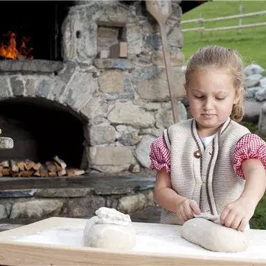 A little girl is working on a piece of dough on a wooden board. In the background, there is a stone oven and a green meadow.