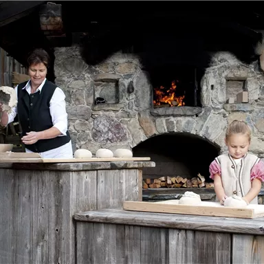 A woman and a girl are baking in a traditional stone oven kitchen. They are working at a wooden table with loaves of bread.