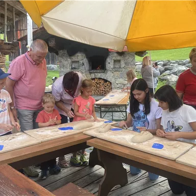 A group of children and adults is preparing dough at large tables outdoors. In the background, there are wooden ovens and a green meadow visible.
