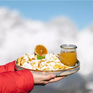 A plate of freshly made Kaiserschmarrn, decorated with powdered sugar and a dried orange slice. Next to it is a glass of fruit juice, set against an impressive mountain backdrop.