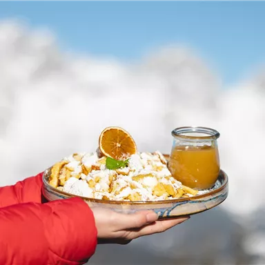 A plate with delicious, fried dough pieces and a slice of orange, served with a glass of juice. In the background, snow-covered mountains can be seen.