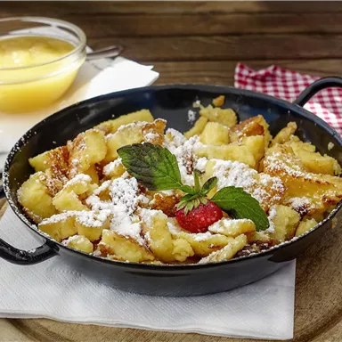 A delicious Kaiserschmarrn, served in a pan, with powdered sugar, fresh berries, and mint. Accompanied by a bowl of applesauce.