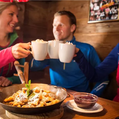 Drei Personen stoßen mit Tassen an und genießen eine Mahlzeit in einer gemütlichen Hütte. Auf dem Tisch liegt eine Schüssel mit einem leckeren Dessert.