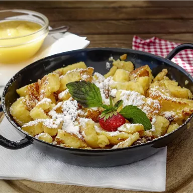 Ein köstlicher Kaiserschmarrn in einer schwarzen Pfanne, garniert mit Puderzucker und einer Frischeminze. Daneben befindet sich eine Schüssel mit Apfelmus.