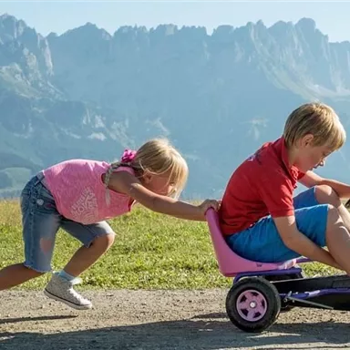 A boy is riding on a small vehicle while a girl pushes him from behind. The scene takes place against an impressive mountain backdrop.