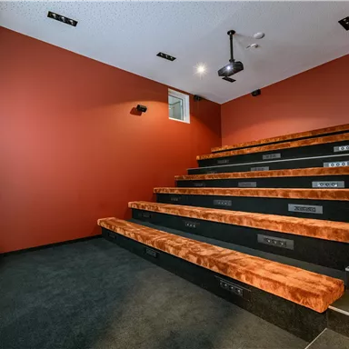 A modern cinema room with tiered seating in orange and a red wall. On the wall, there is a projector and a window that lets natural light in.