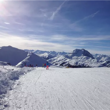A snow-covered mountain landscape under a clear blue sky. Skiers enjoy the slopes in the distance.