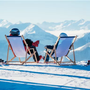 Two people are sitting relaxed on deck chairs in the mountains. In the background, snow-capped peaks and a clear sky can be seen.