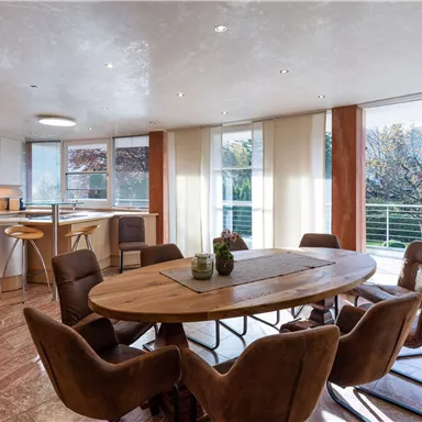 Modern kitchen-living area with a large dining table and cozy chairs. Plenty of daylight through large windows creates an inviting atmosphere.