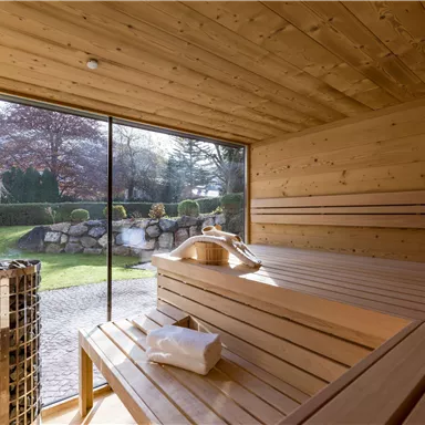 A modern wooden sauna interior with benches and a large window. Outside, trees and a well-maintained garden are visible.
