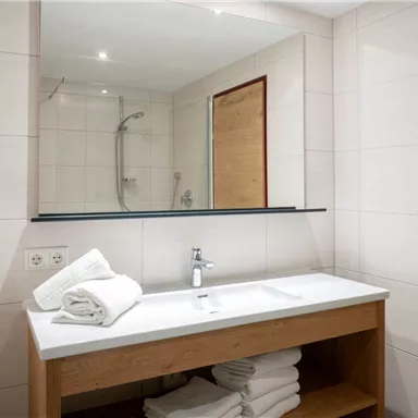 A modern bathroom with a sink, a large mirror, and fresh towels. The walls are brightly tiled, and there is a shower in the background.