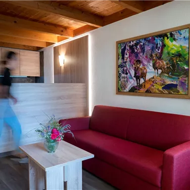 A cozy living room with a red sofa and a wooden table. A colorful painting hangs on the wall, while a person walks by.