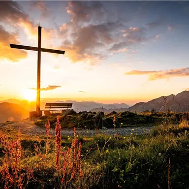 A cross silhouette against a stunning sunset over the mountains. Green meadows and flowers complement the peaceful scene.