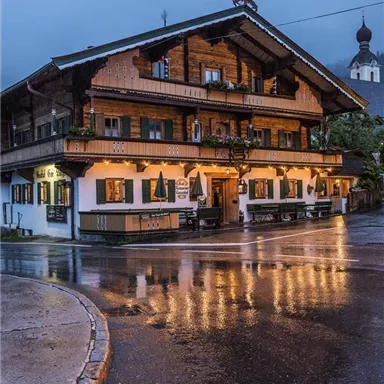 A traditional farmhouse with a wooden facade, illuminated at dusk. Puddles on the street reflect the warm lights of the house.