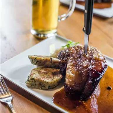 A delicious piece of meat with a golden brown crust rests on a plate, surrounded by a sauce. Next to it are two servings of mashed potatoes and a glass of beer in the background.