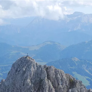 An impressive mountain with rugged rocks and a wide view of the surrounding mountains. The sky is slightly cloudy and the landscape is green and hilly.