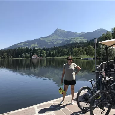 A tranquil lake surrounded by mountains and dense forest. In the foreground, there is a person with a yellow flotation toy and bicycles.