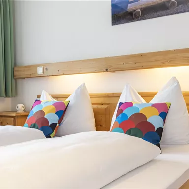 A cozy room with two single beds and colorful pillows. The warm wood look and the green curtains create an inviting atmosphere.