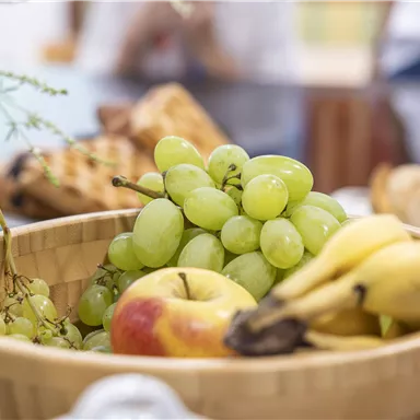 A wooden basket with fresh fruit, including green grapes, an apple, and bananas. In the background, more groceries can be seen.