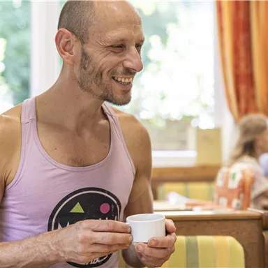 A smiling man is holding a cup in a bright room with a cozy atmosphere. In the background, other people can be seen at the table.