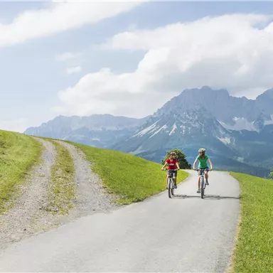 Two cyclists ride on a path through a green landscape. In the background, majestic mountains and a blue sky can be seen.