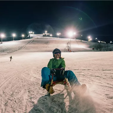 A boy is riding a sled down the illuminated slope. In the background, other winter sports enthusiasts and the snow-covered landscape can be seen.